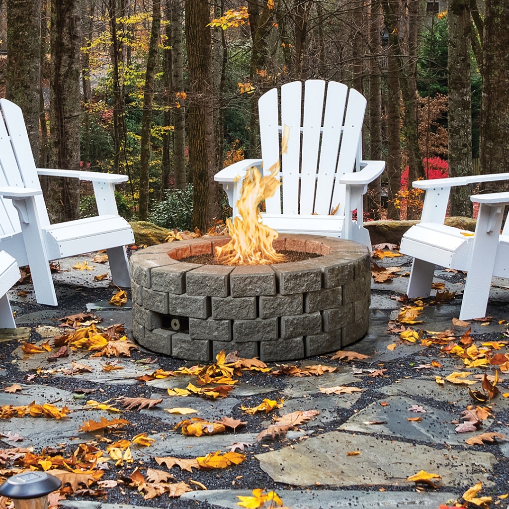 Round stone fire pit with bright flames, surrounded by white Adirondack chairs on a stone patio in autumn leaves.