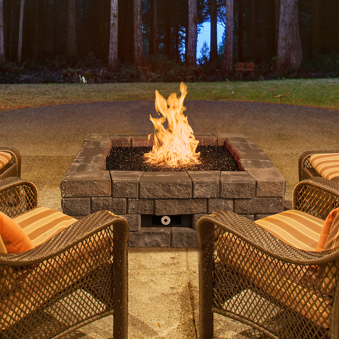 Square brick fire pit with tall flames, surrounded by wicker chairs in a backyard at dusk by a forest edge.