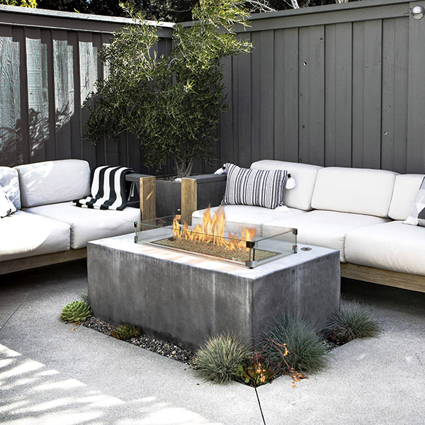 Outdoor lounge with a rectangular concrete fire pit and glass guard, surrounded by white sectional sofas and striped cushions in a fenced backyard.