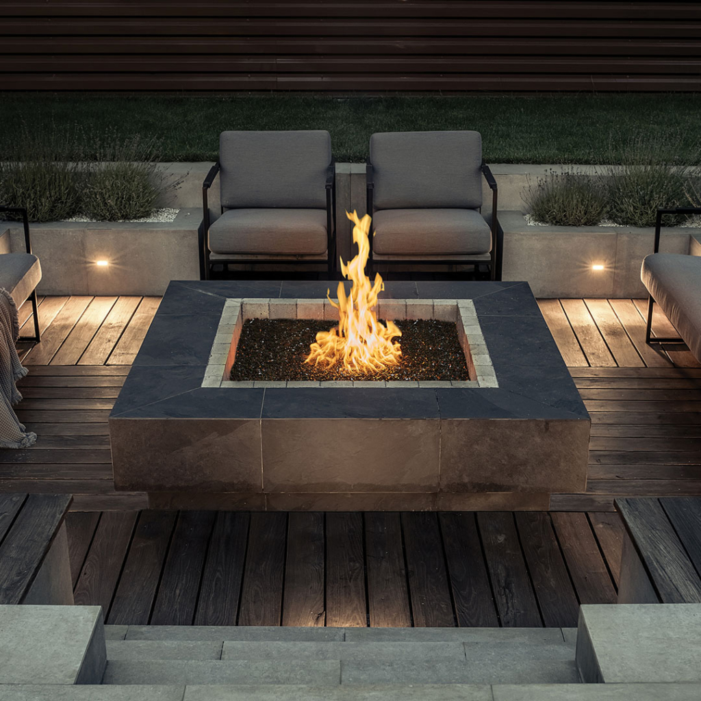 Modern outdoor seating area with a square fire pit at center, flames rising from dark gravel surrounded by stone tiles and wooden deck.