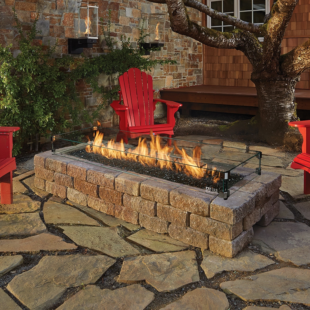 Backyard stone fire pit with a glass guard, flames leaping, surrounded by red Adirondack chairs on a stone patio.