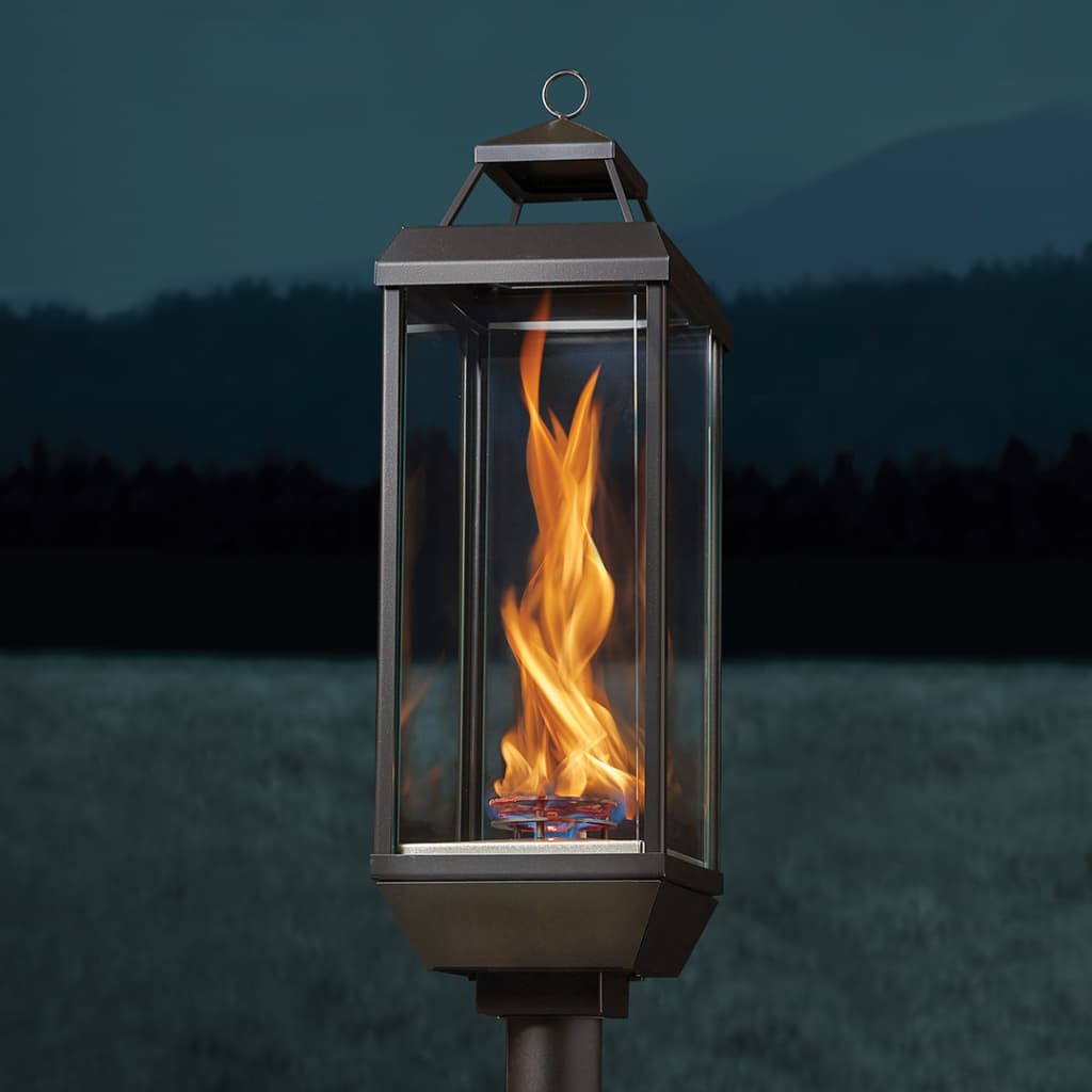 Outdoor metal lantern on a post with a bright flickering flame at dusk by a field/pond.