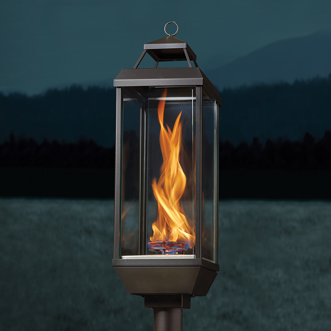 Outdoor metal lantern on a post with a bright flickering flame at dusk by a field/pond.