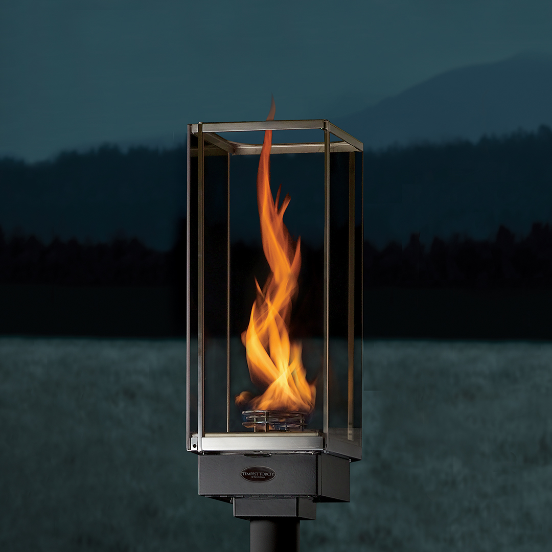 Outdoor metal torch with enclosed flame burning inside a glass frame on a post, against a dark lake and hills at dusk.