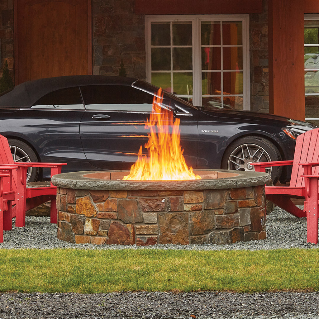 Stone fire pit with bright flames surrounded by red Adirondack chairs on a gravel patio in front of a house with a black car in the background.