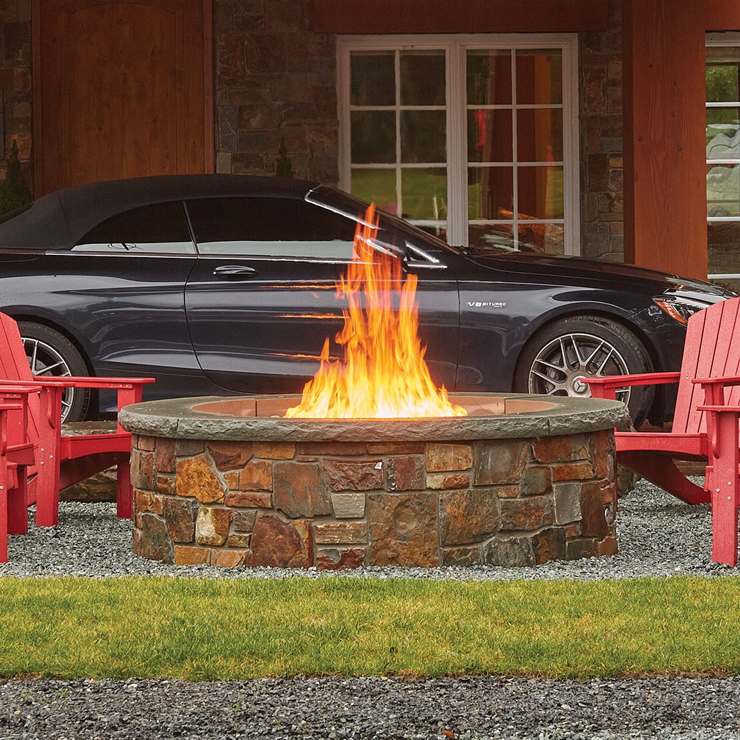 VF47_Install_Sq Stone fire pit with bright flames surrounded by red Adirondack chairs on a gravel patio in front of a house with a black car in the background.