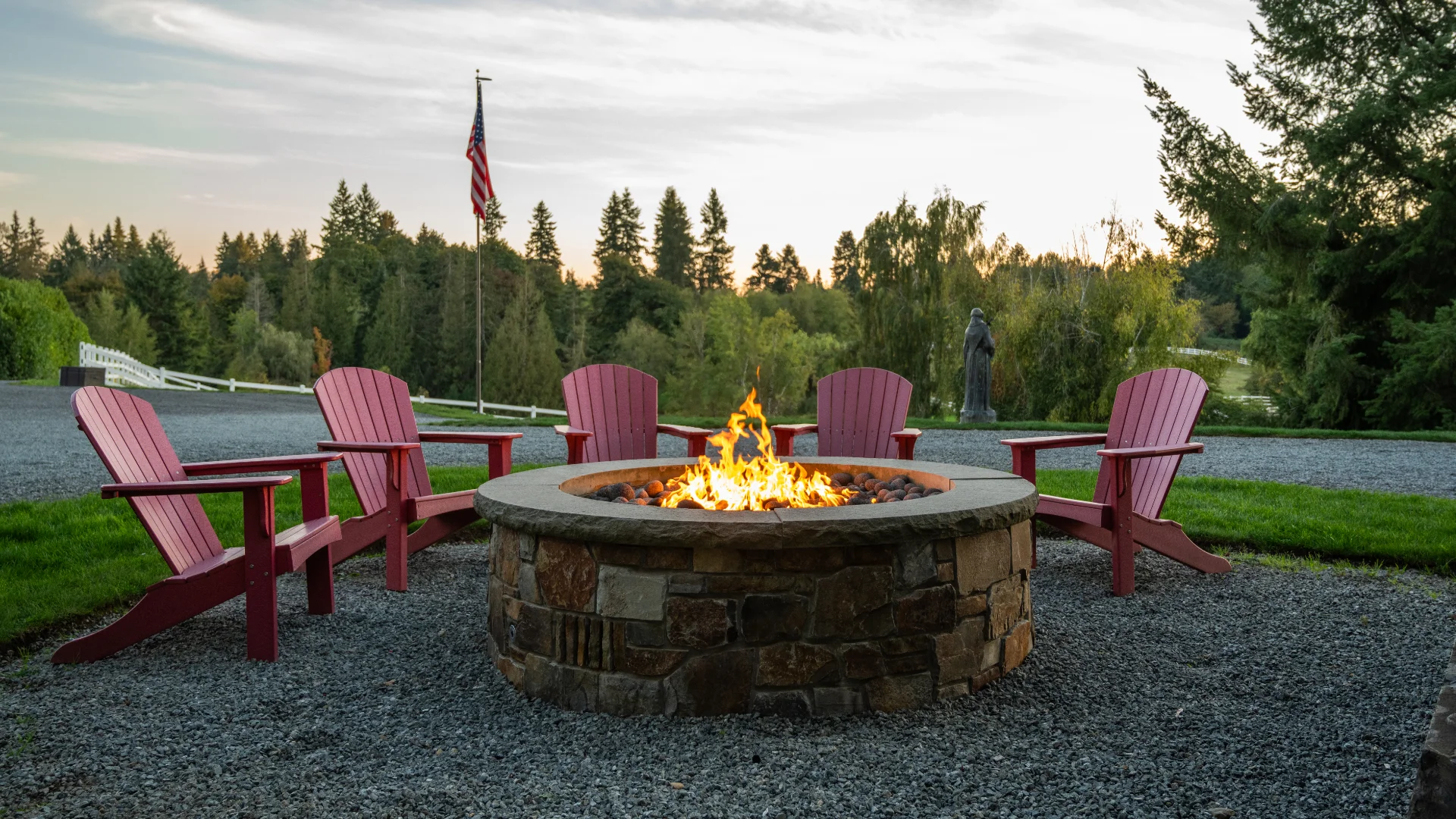 Circular stone fire pit surrounded by six red Adirondack chairs on gravel, with a flagpole and trees in the background at dusk,