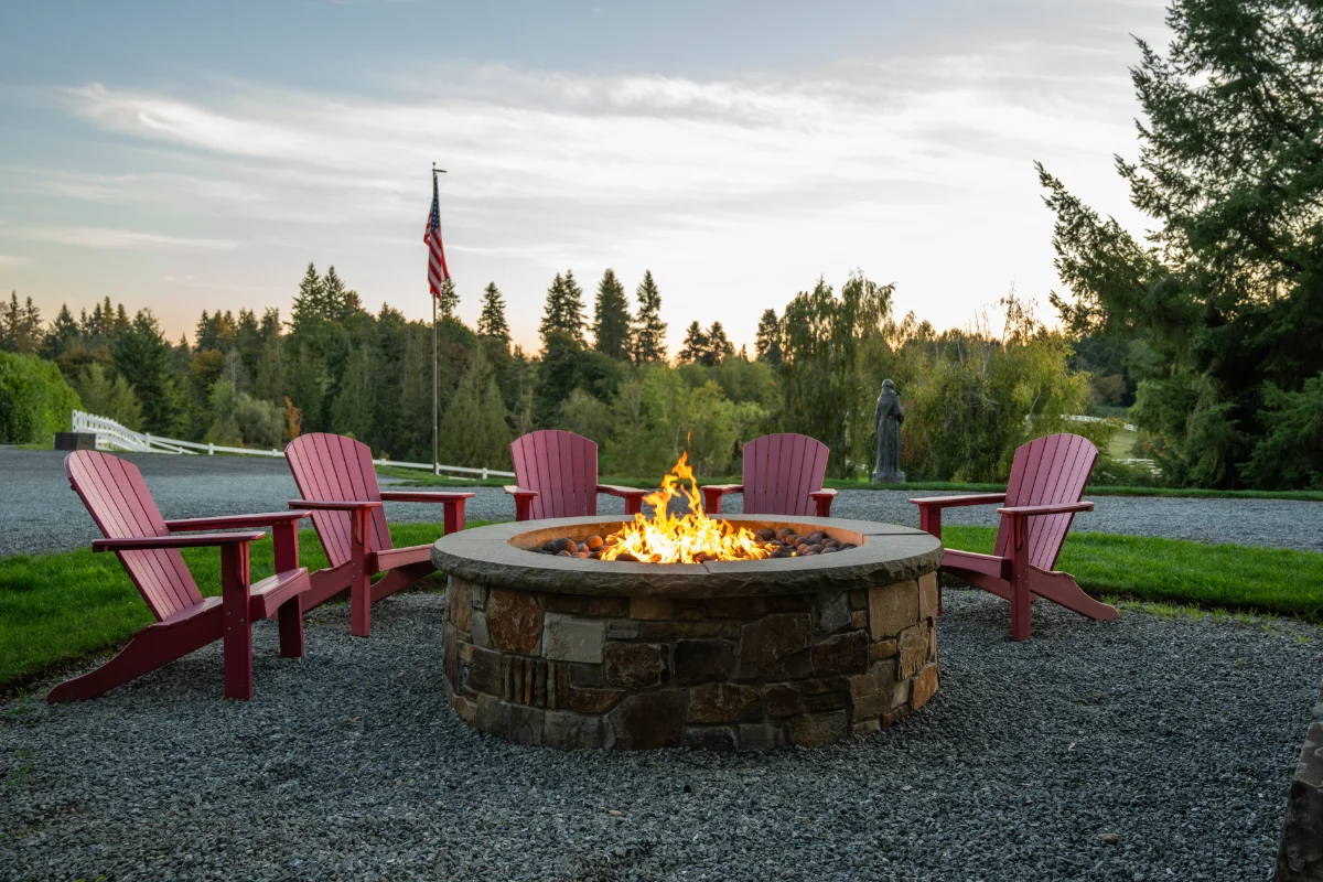 Circular stone fire pit surrounded by six pink Adirondack chairs around gravel, with a flagpole and trees at sunset.
