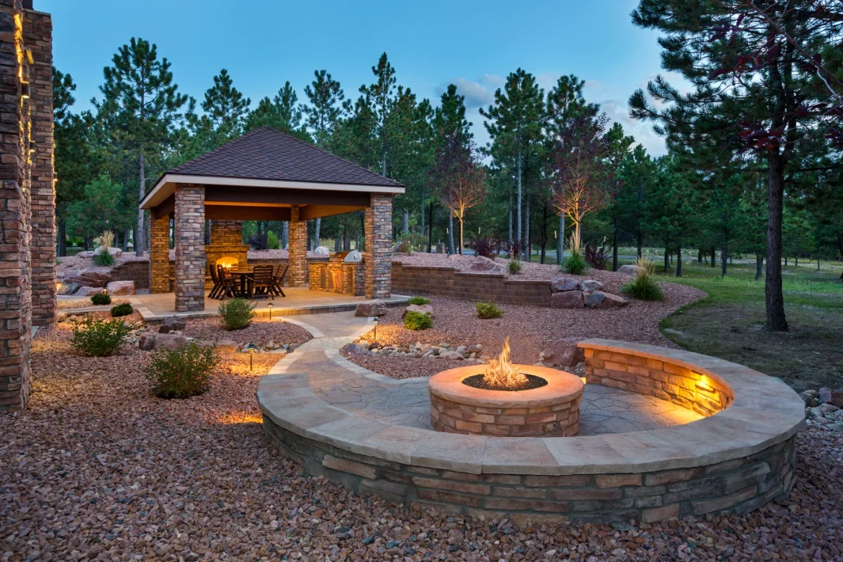 Circular stone fire pit with flame, built-in curved seating, and gravel landscaping in a wooded backyard.