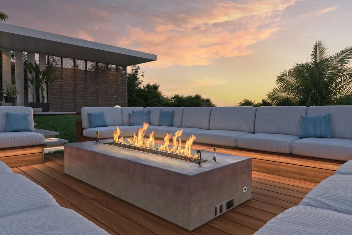 Outdoor modern lounge area around a rectangular concrete fire pit, white cushioned seating, blue throw pillows, at sunset.