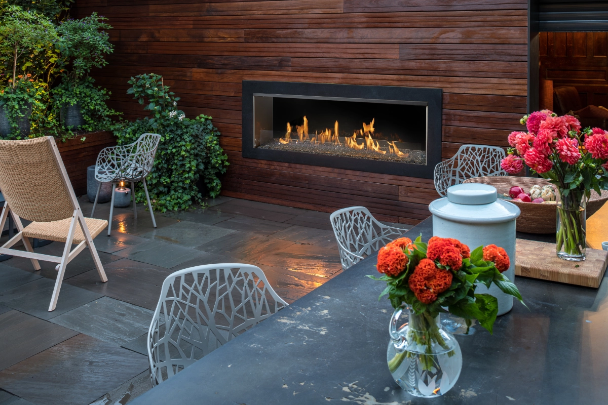 Outdoor patio with a long, rectangular gas fireplace set into a wood-paneled wall, surrounded by metal seating and a table with orange flowers in a vase.