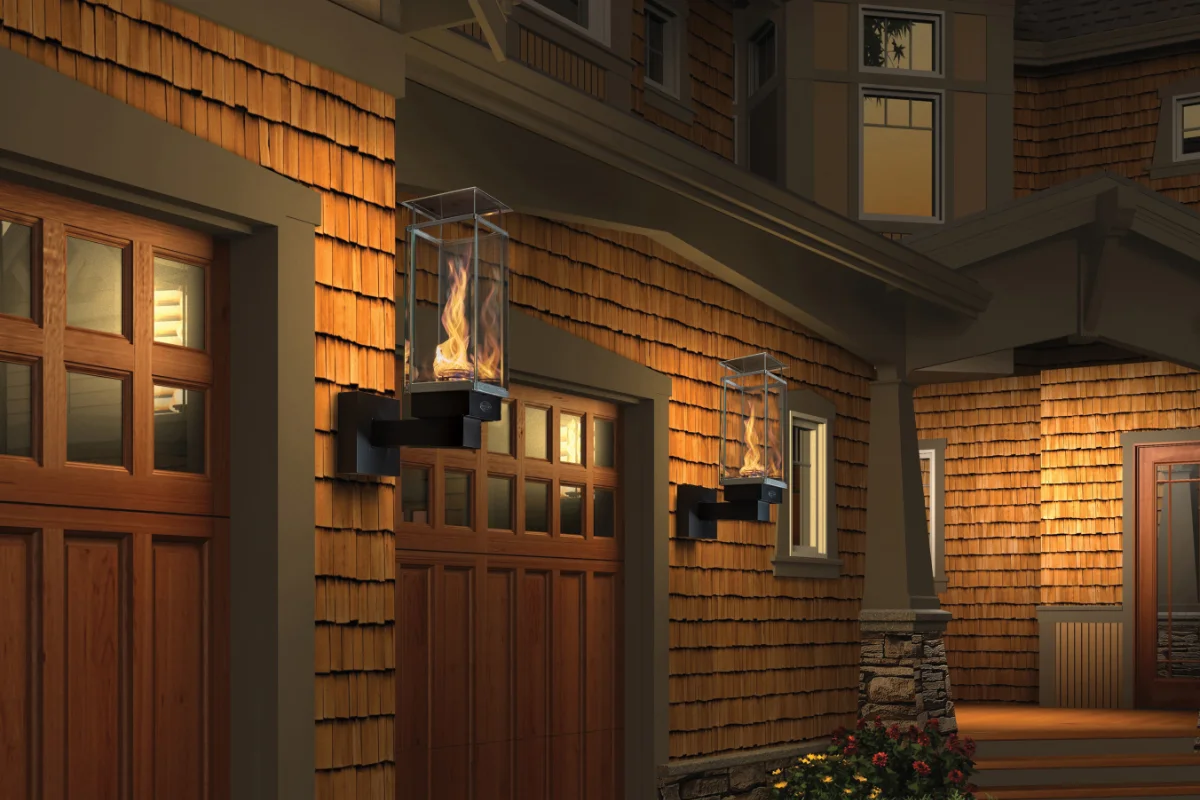 Exterior of a wooden-clad home at dusk with three wall-mounted glass lanterns burning beside garage doors.